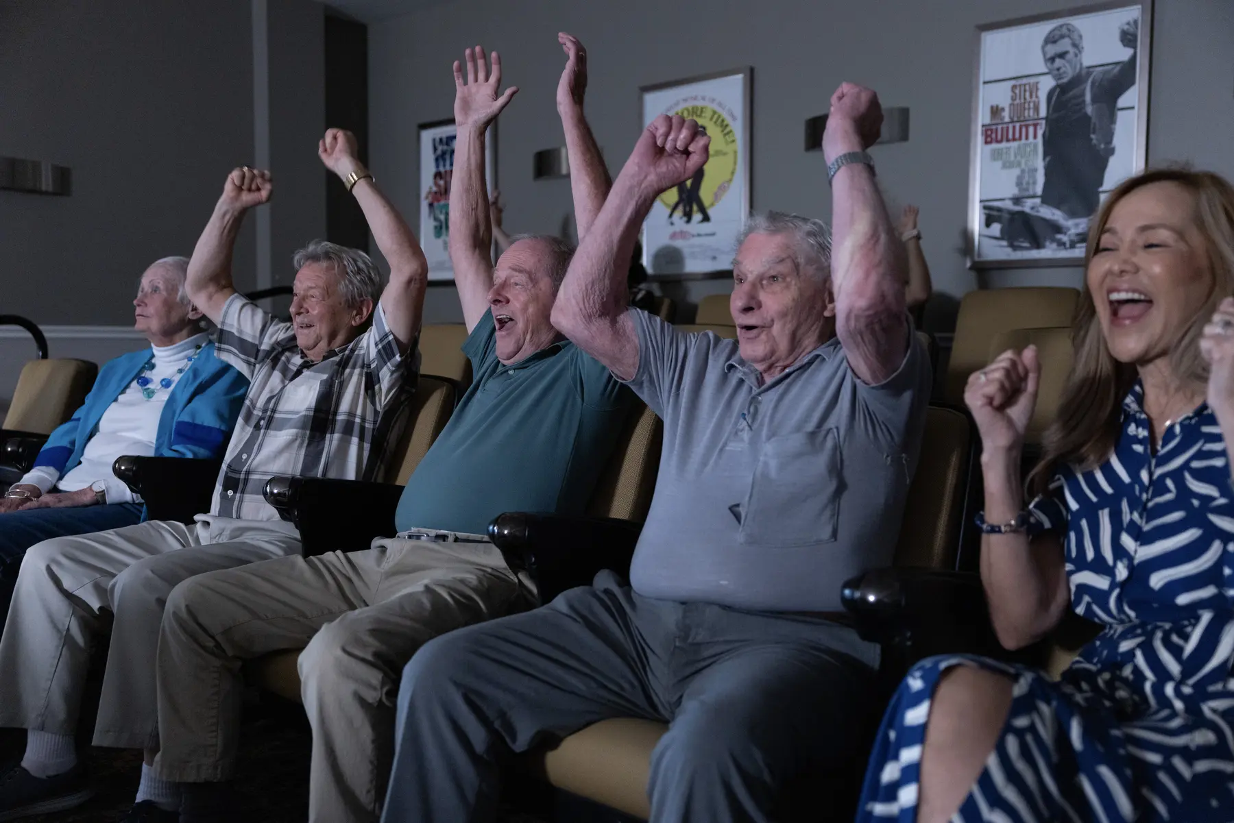 Seniors sitting in a cinema looking towards a movie screen, cheering with hands in the air.
