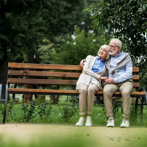 smiling senior couple embracing and holding hands while resting on wooden bench in park