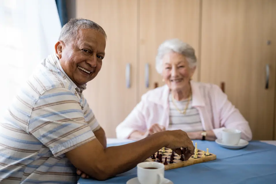Senior man and senior woman is looking at the viewer while having a coffee and playing a chess / Un homme et une femme âgés regardent le spectateur tout en prenant un café et en jouant aux échecs