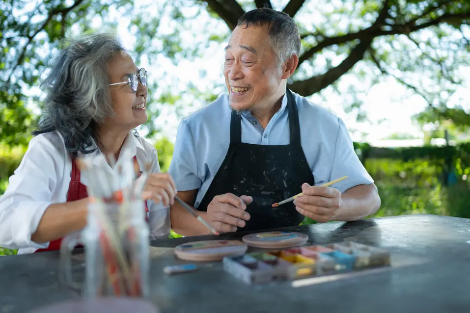 Seniorasiancouplepaintingpotterytogetheratatableoutside.