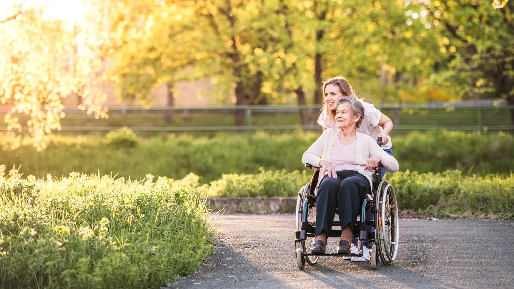 Elderly grandmother in wheelchair with an adult granddaughter outside in spring nature / Une grand-mère âgée en fauteuil roulant avec sa petite-fille adulte à l'extérieur, dans la nature printanière