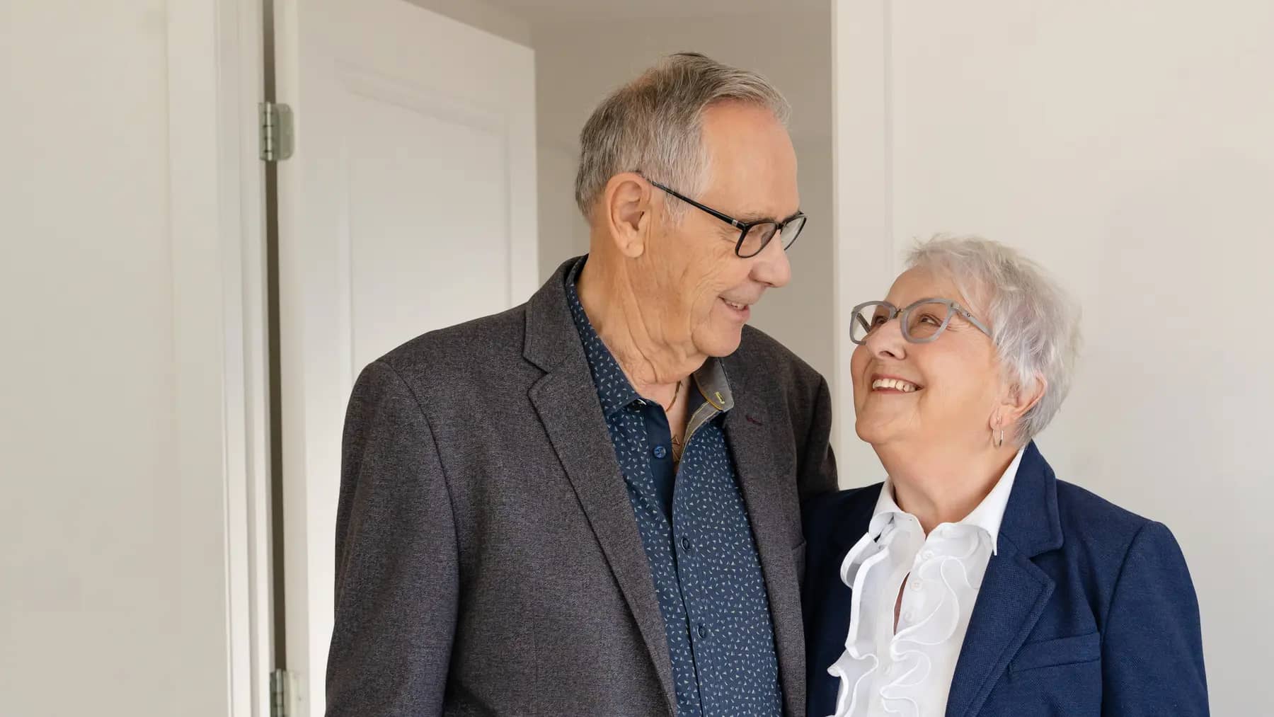 Happy senior couple hugging each other and standing in a suite / Couple âgé heureux s'étreignant et debout dans une suite