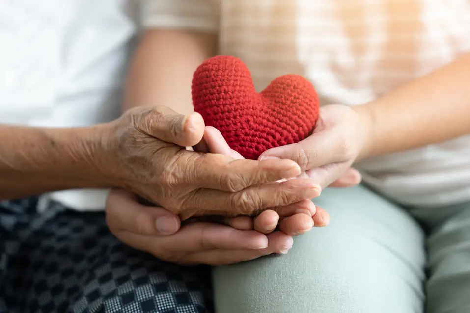 Young and senior woman holding each other hands and red yarn heart shape togetherness concept. Elderly care and protection with love from grandchild.