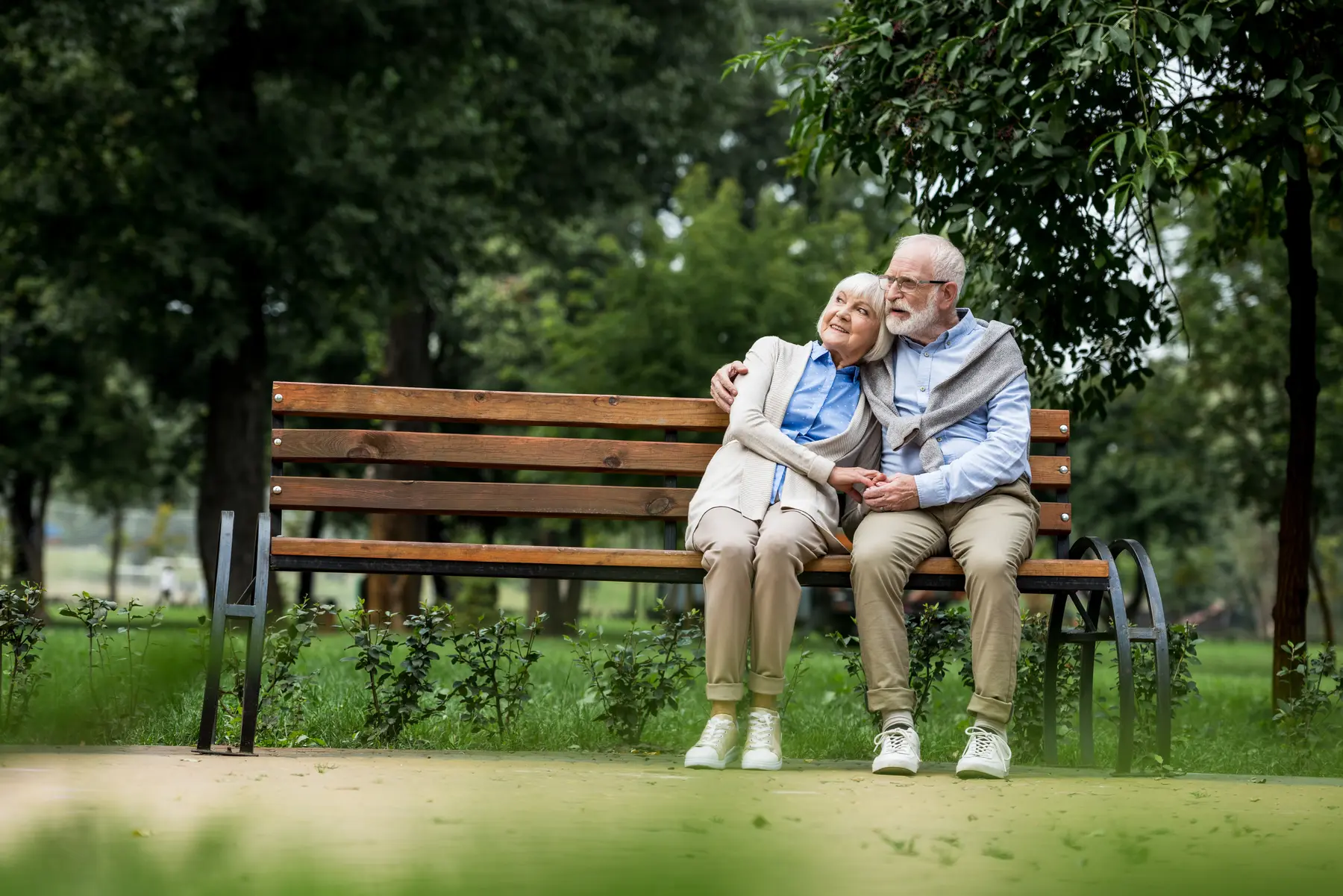 smiling senior couple embracing and holding hands while resting on wooden bench in park / Couple âgé souriant s'embrassant et se tenant la main, assis sur un banc en bois dans un parc