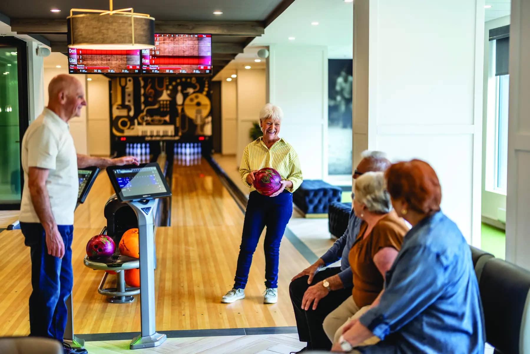 Seniorsbowlingatasmallindooralley.Thecurrentbowlerisawomanholdingabowlingballinherhand,smilingandstaringbackatherfriendswhoaresittingwatching.