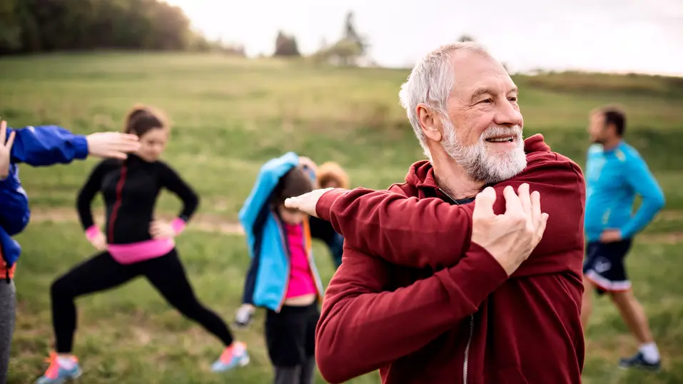Senior man in wearing red hood, stretching out on a grass field / Homme âgé portant une cagoule rouge, s'étirant sur un champ d'herbe