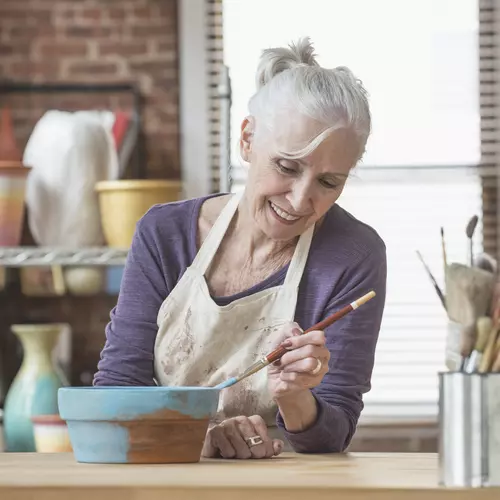 Senior woman in a studio painting a pot of cloy with a brush / Femme âgée dans un atelier peignant un pot de cloy au pinceau