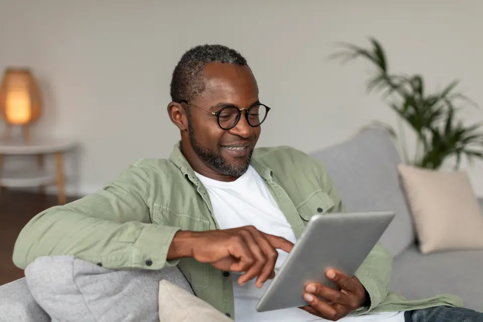 Older adult using a tablet while relaxing on a sofa at home. / Personne âgée utilisant une tablette tout en se détendant sur un canapé à la maison.
