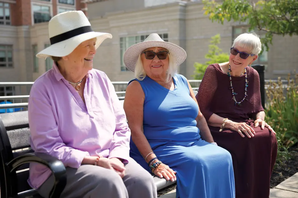 Three senior ladies wearing colourful dresses are sitting on a bench outdoors