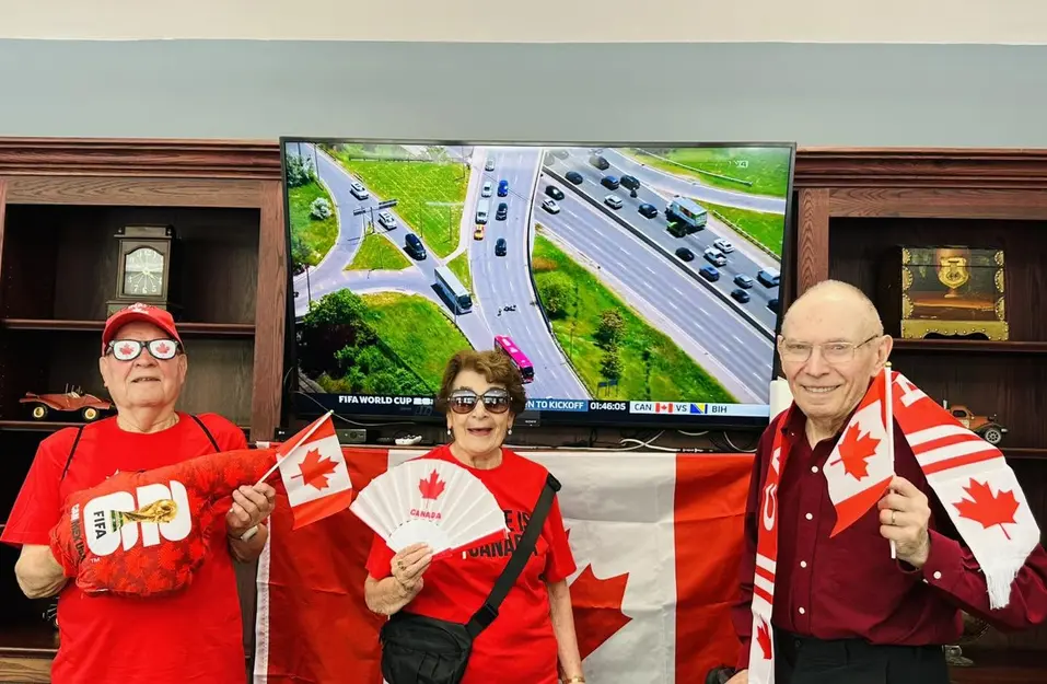 Residents holding Canadian flags and cheering for a televised soccer match in a retirement residence / Résidents tenant des drapeaux canadiens et encourageant un match de soccer télévisé dans une résidence pour aînés