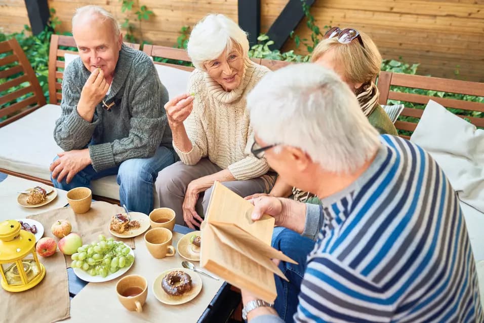Group of seniors sitting in outdoor patio enjoying coffee cake and fruit while reading a book