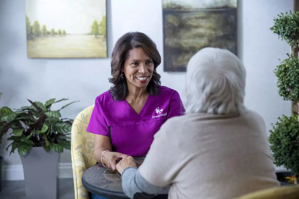 Chartwell care support staff sitting with the senior resident, holding hands smiling and having cheerful conversation. / Le personnel soignant de Chartwell assis auprès d'un résident âgé, lui tenant la main, souriant et discutant joyeusement.