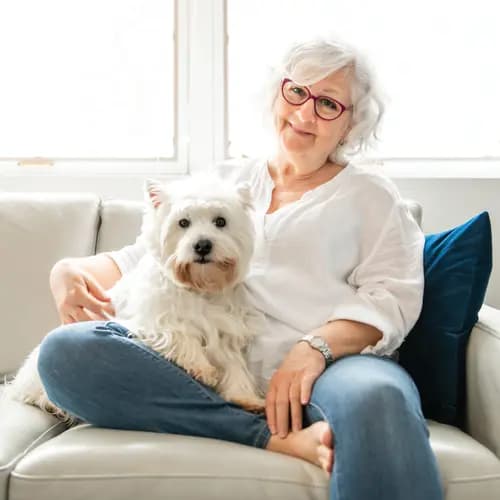 Senior woman is sitting on her couch along with her white dog / Une femme âgée est assise sur son canapé avec son chien blanc.