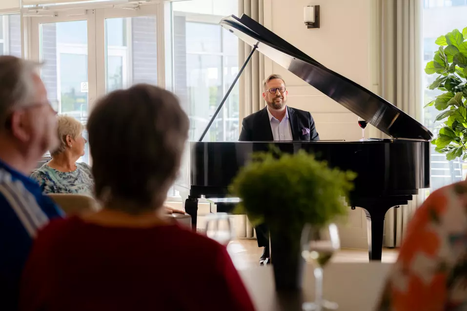 Seniors gathered and listening to a man playing a grand piano / Des personnes âgées se rassemblent et écoutent un homme jouant du piano à queue