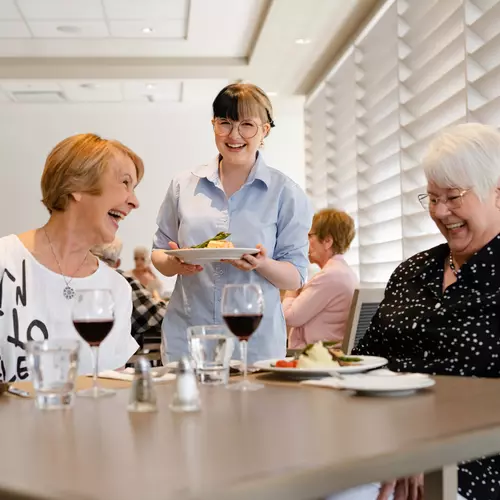Server at a Chartwell residence is serving a dinner to smiling senior women / Un serveur d'une résidence Chartwell sert un dîner à des femmes âgées souriantes